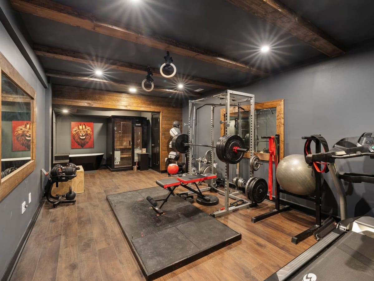 A home gym room with dark walls and exposed beams: a treadmill in the foreground, a squat rack with a loaded barbell, adjustable dumbbells and a rubber lifting platform — Belt & Bike, home treadmills & exercise bikes