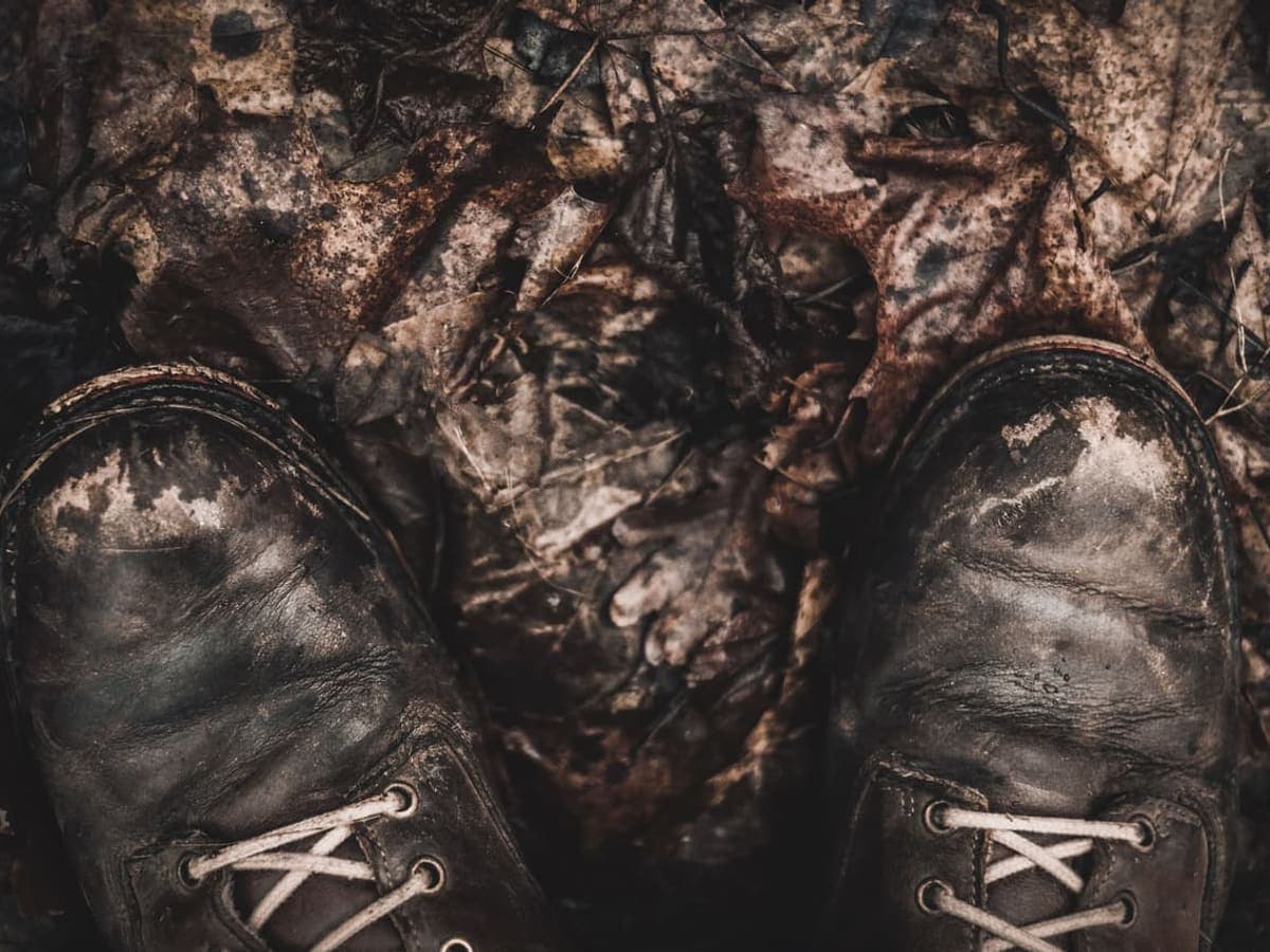 Looking down at a pair of mud-caked brown leather boots standing in wet autumn leaf litter — Bog & Briar, hunting boots & snake boots