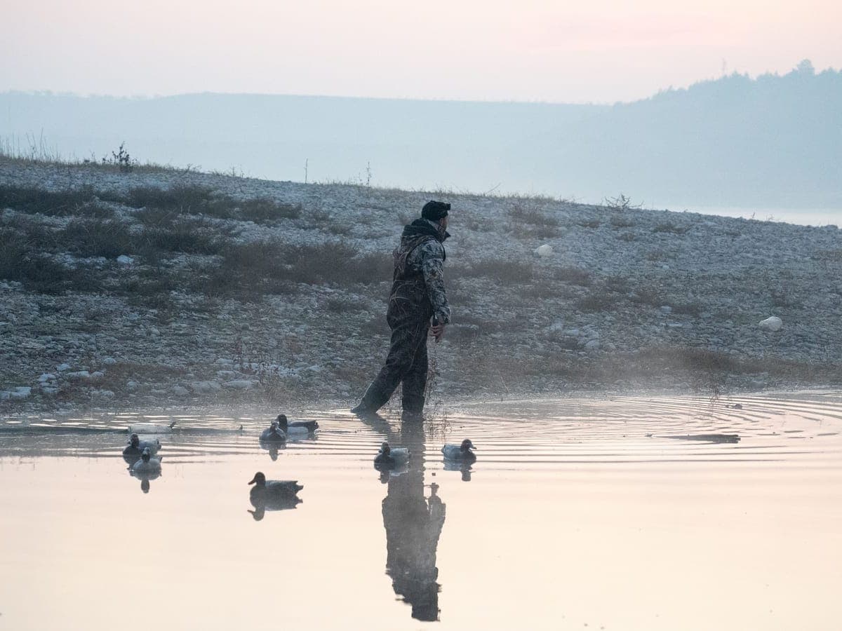 A hunter in camouflage waders setting a spread of duck decoys on misty water at dawn — Decoy & Drake, waterfowl hunting gear