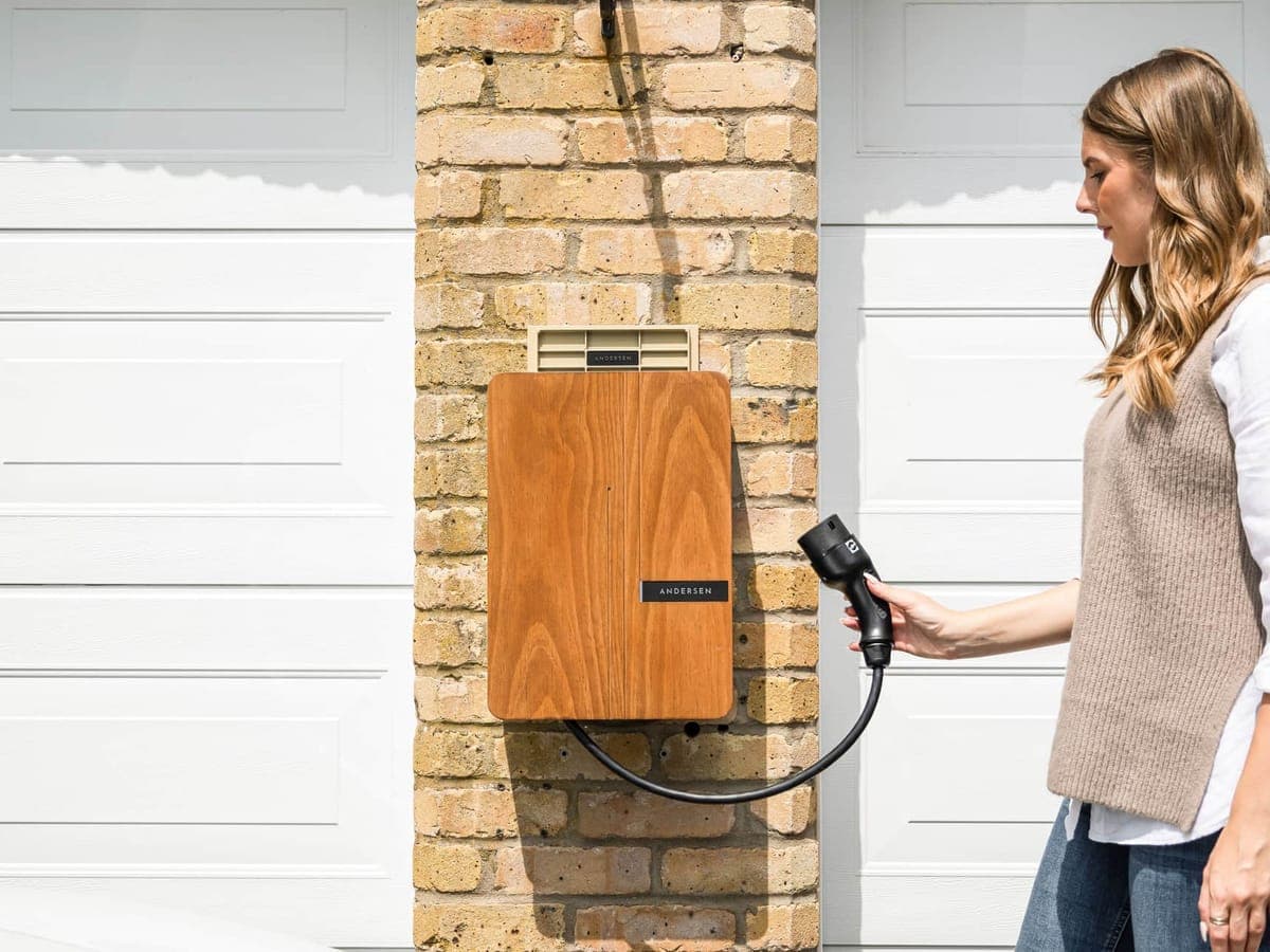 A woman holding the connector of a wood-fronted home EV charger mounted on a brick pillar between two white garage doors — EV & Charger, home ev charging