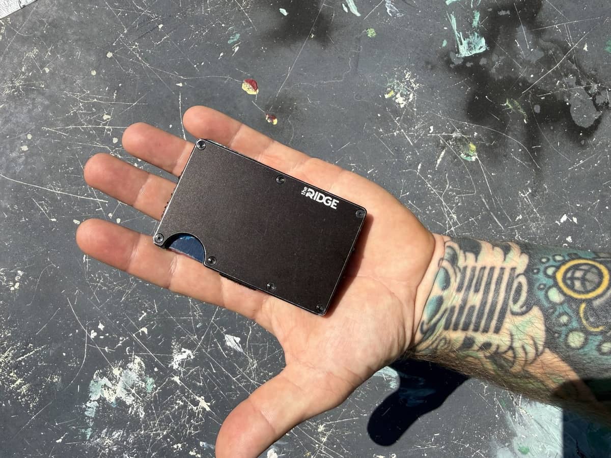 A slim metal everyday-carry wallet held flat in an open, tattooed hand over a scratched workbench — Every Dad Carry, everyday carry & dad gear