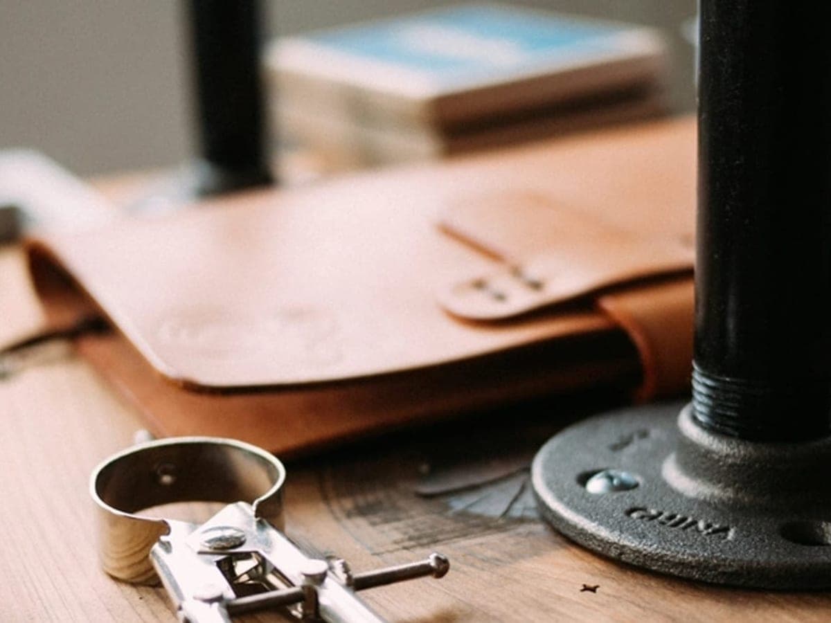 A folded piece of tan vegetable-tanned leather on a wooden workbench beside a metal clamp and a cast iron pipe flange — Forge & Forever, boots, leather & buy-it-for-life gear