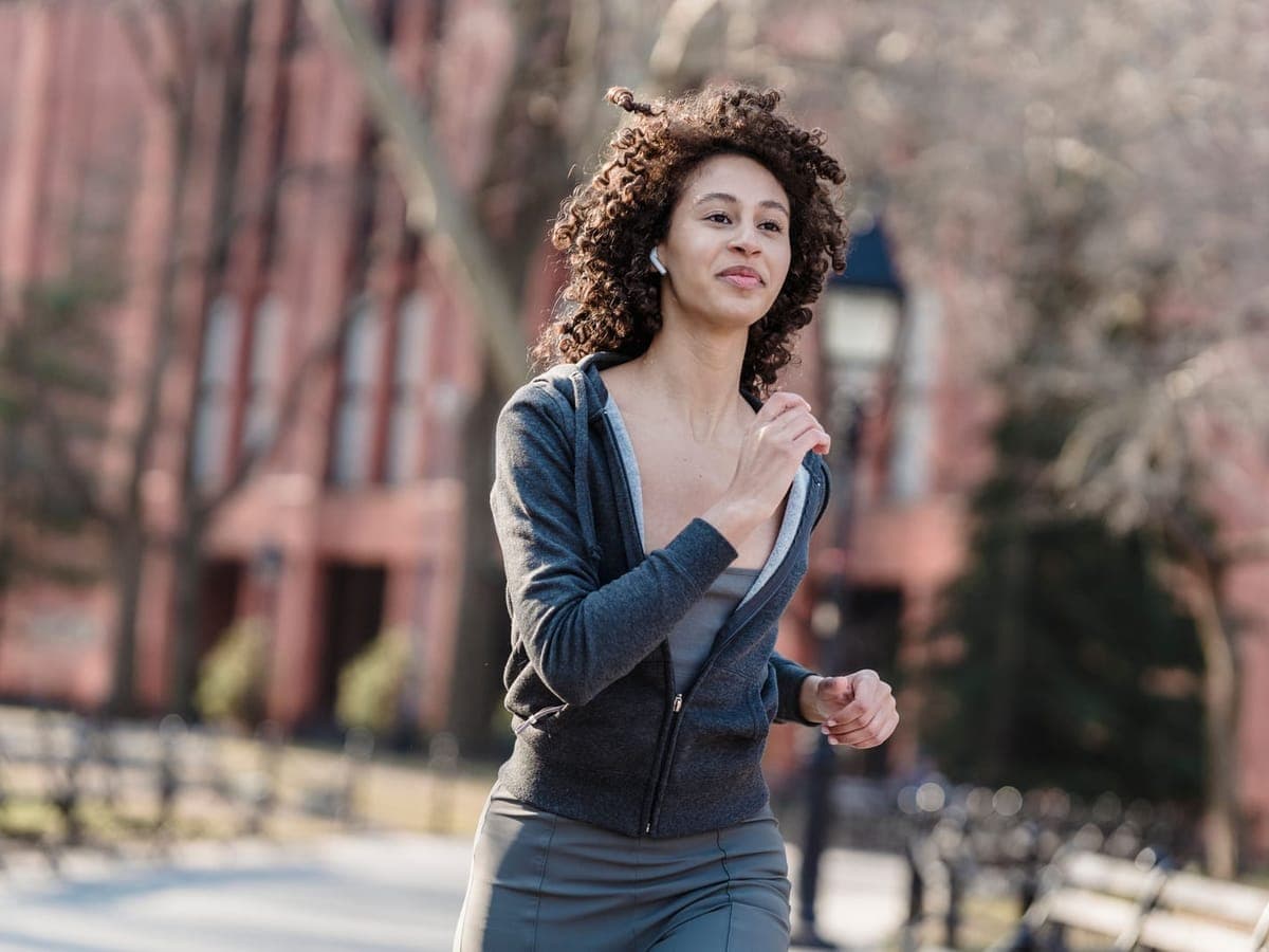 A woman with curly hair jogging past red brick buildings in a city park, wearing a small white earbud — Openear Club, open-ear & workout earbuds