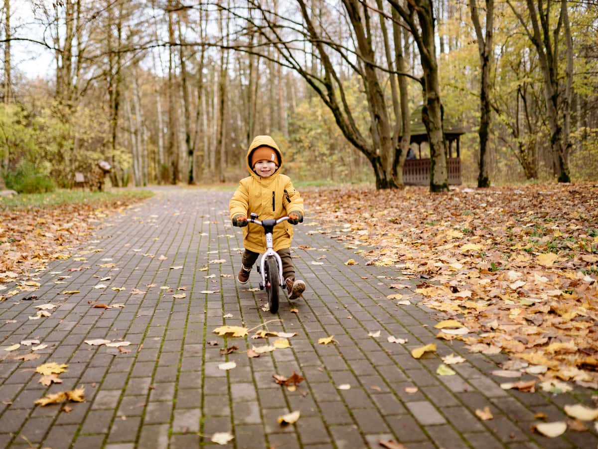 A small child in a yellow jacket riding a balance bike on a leafy path — Pedal & Pint, kids bikes & balance bikes