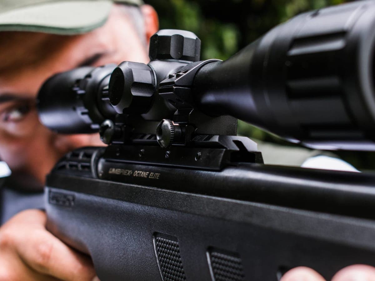 A hunter in a cap aiming a scoped rifle, seen close over the scope and rail — Sounder & Scope, hog & night hunting gear